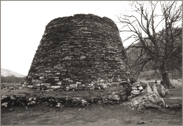 The exterior of the broch showing the surrounding wall. Two people can be seen at the base of the tree on the right, giving some idea of the scale
